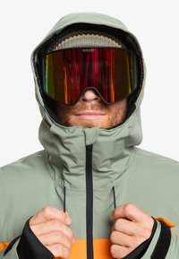 Chaqueta de esquí verde y naranja con cuello alto, que cuenta con una capucha ajustable y acentos negros. Lleva puesta unas grandes gafas de esquí espejadas.