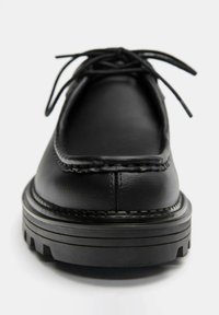 Sort lædersko med rundet tå, syninger ved kanterne, flade såler og snørebånd foran. Har en tekstureret overflade og et minimalistisk design.