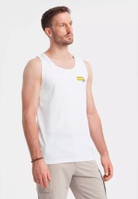 Bílý bavlněný top na ramínka s kulatým výstřihem a logo detailem v žluté a zelené barvě na levé části hrudi. Model má na sobě khaki kalhoty.