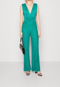 Mono de lentejuelas en teal con escote en V profundo, diseño plisado, piernas amplias y cintura ajustada. Combinado con un bolso negro y sandalias de dedo abierto.