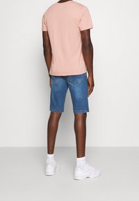 T-shirt en coton rose clair, shorts en denim bleu avec ourlets retroussés, portés avec des chaussures de sport blanches et des chaussettes mi-mollet, vus de dos.