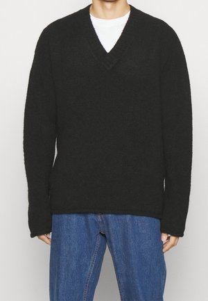 Mand iført sort, loddent V-hals sweater over hvid skjorte og blå jeans, stående mod en ensfarvet lys baggrund.