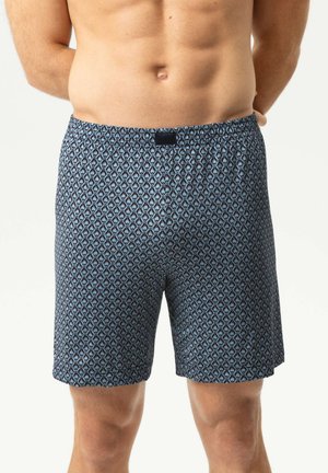 Erwachsener Mann trägt blaue gemusterte Boxershorts, steht mit den Händen hinter dem Rücken vor einem einfarbigen Hintergrund.