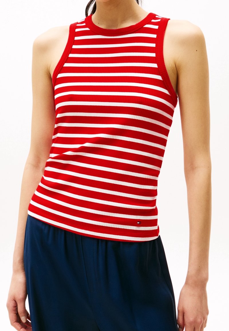 Femme portant un haut sans manches à rayures horizontales rouges et blanches associé à un pantalon bleu foncé, debout devant un fond blanc uni.
