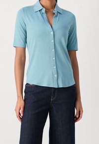 Blusa azzurro chiaro a maniche corte con colletto e chiusura frontale a bottoni, realizzata in tessuto elasticizzato, abbinata a pantaloni in denim scuro.
