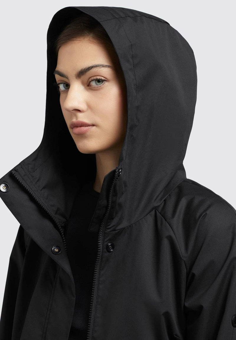 Veste noire imperméable avec capuche, dotée d'une fermeture éclair et de boutons-pression, texture lisse et design minimaliste.