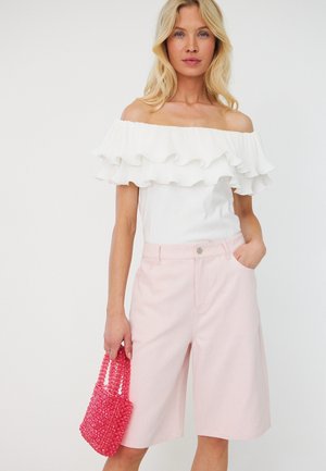 Femme portant un haut blanc à volants et épaules dénudées ainsi qu’un short rose clair à hauteur des genoux, tenant un petit sac à main rose perlé dans une main.