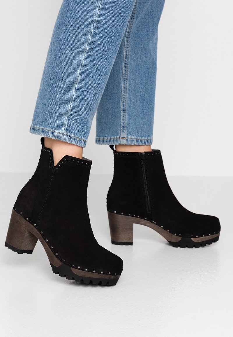 Bottines en daim noir avec talons blocs en bois, clous métalliques le long de la semelle, portées avec un jean court bleu clair sur un sol blanc.