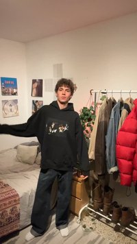 Jeune personne portant un sweat à capuche noir et un jean, se tient dans la chambre près du lit, portant un portant avec des vestes, des bottes et une plante verte.