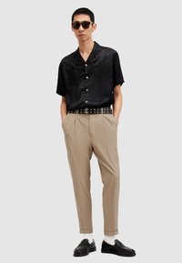 Camisetta nera a maniche corte con ricami, pantaloni beige con pieghe, cintura nera con dettagli ad occhiello, mocassini neri e calze bianche. Occhiali da sole indossati.
