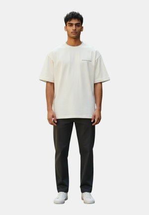 Giovane uomo in piedi di fronte a sé, indossa una maglietta bianca larga, pantaloni neri e scarpe da ginnastica bianche su uno sfondo semplice.