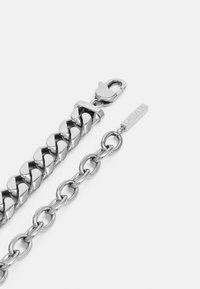 Pulsera de cadena de plata con eslabones entrelazados, un cierre de langosta y una etiqueta con la inscripción "Lacoste". Acabado de metal suave y brillante.