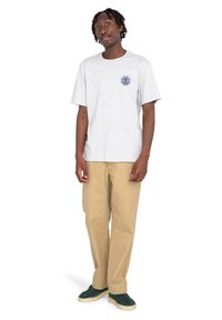 Sivá bavlnená tričko s okrúhlym golierom, predným logom, v kombinácii s béžovými nohavicami a zelenými teniskami. Model stojí vzpriamene, ruky má uvoľnené.