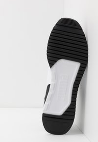 Talpa de sneaker alb-negru cu o suprafață din cauciuc texturat, având un logo alb proeminent în partea de mijloc și striuri orizontale pentru aderență.