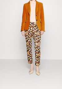 En orange fløjl blazer over en lyserød top, parret med brune bukser med et farverigt kyllingemønster. Beige sko fuldender looket.