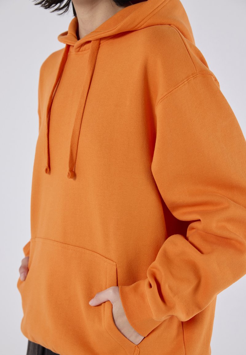 Sweatshirt à capuche orange en tissu doux, avec une poche avant et un cordon de serrage. Les poignets et l'ourlet sont côtelés pour un look ajusté.
