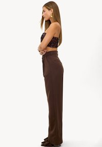 Pantalon marron taille haute, tissu lisse, avec une poche latérale, associé à un haut bustier sombre à sequins. Le mannequin est de profil.