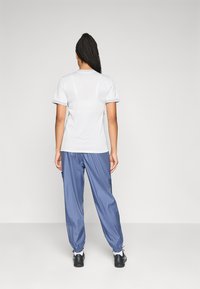 Une personne ayant de longs cheveux tressés se tient face à un mur sobre, portant une chemise blanche à manches courtes, un pantalon bleu ample et des chaussures noires.