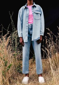 Vaaleansininen farkkupaita vaaleanpunaisen pyöreäkaulaiseen paidan päällä, yhdistettynä korkeavyötäröisiin vaaleansinisiin farkkuihin ja valkoisiin puukenkään, tummalla taustalla.