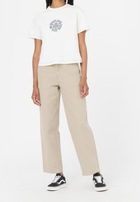 Vit kortärmad t-shirt med en grafisk logotyp, kombinerad med beiga löst sittande byxor och svarta sneakers med vita detaljer.