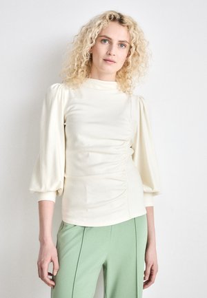 Elfenbeinfarbener Pullover mit Puffärmeln, seitlichen Raffung und hohem Ausschnitt. Kombiniert mit hellgrünen, taillierten Hosen. Glatte Textur.