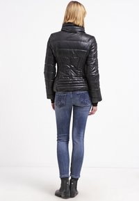 Schwarze Daunenjacke mit hohem Kragen, tailliertem Design und gestepptem Muster; kombiniert mit blauen Jeans, die ein Blumenmuster auf den Gesäßtaschen haben.