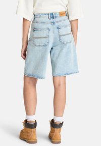 Shorts en denim bleu clair avec une coupe droite, deux poches arrière et une étiquette en cuir à la taille. Associés à des bottes jaunes et des chaussettes blanches.