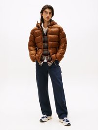 Giacca a imbottitura marrone con cappuccio, indossata sopra una camicia a quadri, abbinata a pantaloni blu navy e sneaker bianche. La giacca ha una texture liscia e lucida.