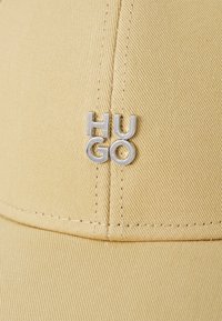 Žltá čiapka vyrobená z textúrovanej látky, na prednej strane so strieborným logom "HUGO". Dizajn obsahuje detailné švy a zakrivený šilt.
