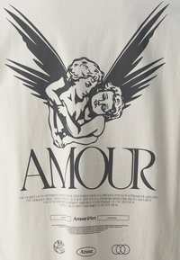 Kremo spalvos marškinėliai su juodu grafiniu dvejais sparnuotais cherubais, besikabinančiais tarpusavyje, ir žodžiu "AMOUR", išryškintai parašytu storomis raidėmis žemiau.