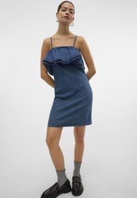 Denim obleka v srednje modri barvi z nabranim ovratnikom, prilegajočo silhueto in naramnicami. Ujemajo se s sivimi nogavicami in črnimi loafers.