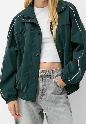 Vrouw met een donkergroen jack met rits en witte biezen, een wit cropped topje en lichtgrijze, hooggetailleerde spijkerbroek, hand in de zak.