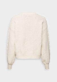 Krémový svetr s texturou a volným střihem. Má spadená ramena, žebrované manžety a tkaný vzor na rukávech.