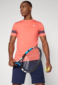 Koral skjorte med korte ermer og marineblå striper på ermene. Modellen holder en tennisracket i den ene hånden og en grønn tennisball i den andre.