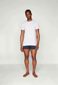 T-shirt blanche à manches courtes avec col rond, associée à un boxer bleu à motifs. Modèle debout pieds nus, mettant en valeur la tenue.
