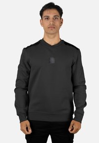 Sudadera negra de tela suave, con cuello redondo, mangas largas y un pequeño parche de logo en el pecho, con detalles texturizados en los hombros.