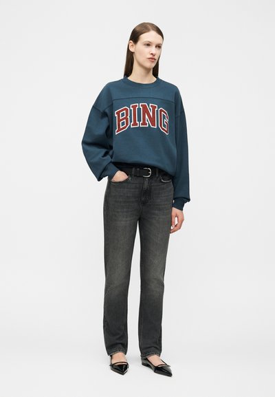 Kvinne stående iført en blå sweatshirt med teksten "BING", svart belte, falmet svarte jeans og svarte sko med spiss tå mot en hvit bakgrunn.