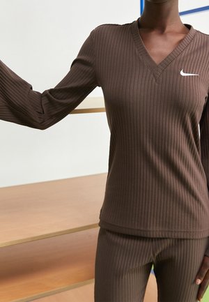 Haut à manches longues marron côtelé avec un décolleté en V et un petit logo Nike blanc sur la poitrine gauche. Tissu doux et design ajusté.