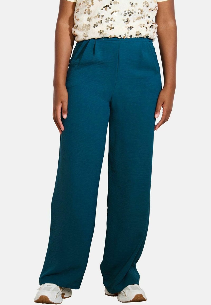 Pantaloni a gamba larga color teal realizzati in un tessuto testurizzato, con vita alta e tasche laterali, abbinati a una maglietta leggera e fantasia e scarpe da ginnastica bianche.