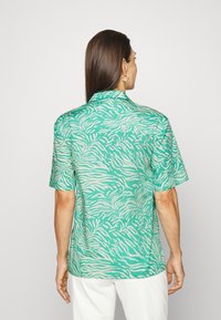Camisa de manga curta em tecido verde com padrão de ondas abstratas brancas, apresentando colarinho e um corte confortável. As costas estão visíveis.