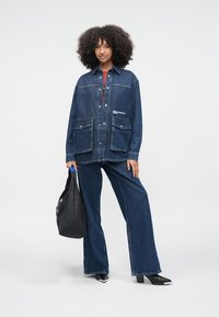 Kurtka dżinsowa w ciemnym niebieskim kolorze z dwoma przednimi kieszeniami i zatrzaskami, noszona z dopasowanymi, szerokimi spodniami dżinsowymi. Czarne buty na obcasie i czarna torebka.