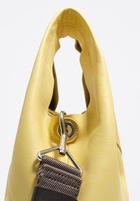 Żółta skórzana torebka z unikalnym uchwytem w kształcie wycięcia, srebrnymi akcentami oraz odczepianym brązowym paskiem z teksturowanej tkaniny.