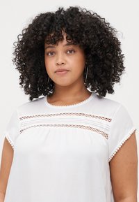 White blouse with short sleeves, featuring lace trim and a textured yoke with horizontal and vertical lines. Smooth fabric, casual design.