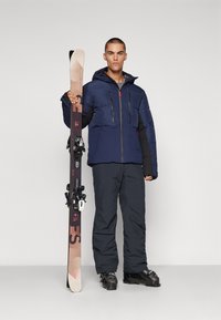 Navy puffer jacket with black accents, black ski pants, and black ski boots. Holds brown and pink skis with black bindings. Simple background.