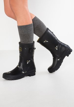 Piernas con calcetines grises y botas de lluvia negras hasta la mitad de la pantorrilla decoradas con patrones de abejas amarillas y blancas sobre un suelo blanco.
