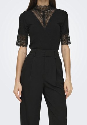 Femme portant un pantalon noir taille haute et un top noir à manches courtes avec des détails en dentelle sur l'encolure et les manches.