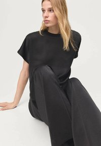 Schwarzes, kurzärmeliges Oberteil kombiniert mit schwarzen, weit geschnittenen Hosen aus glattem Stoff und entspannter Passform, vor neutralem Hintergrund.