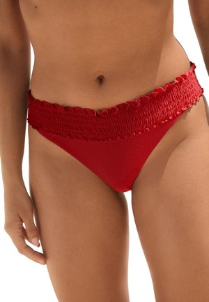 Kvinde iført røde bikinitrusser med en rynket, flæset talje, der viser maven og højre hånd mod en hvid baggrund.