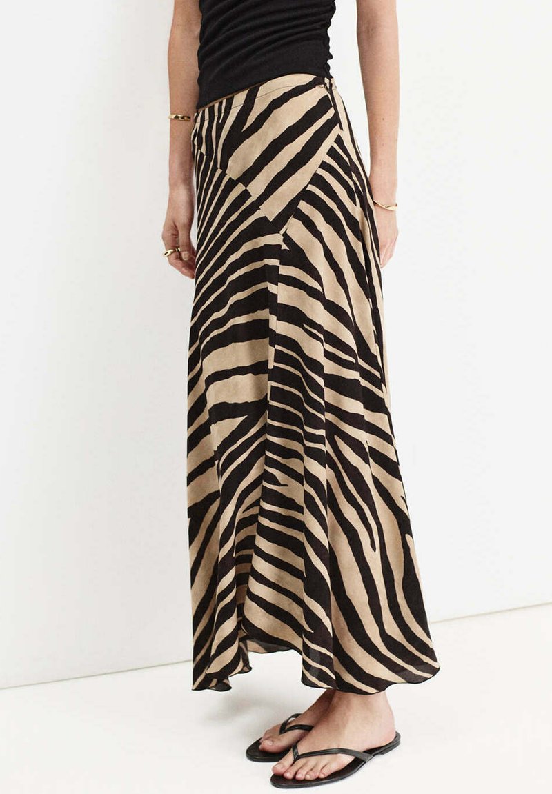Femme portant une jupe longue beige et noire à rayures zébrées, un top noir sans manches, des bracelets dorés et des tongs noires, debout contre un mur blanc.