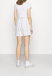 Femme portant un short blanc taille haute à rayures verticales noires, un t-shirt court blanc, et des baskets blanches montantes, debout contre un mur uni.